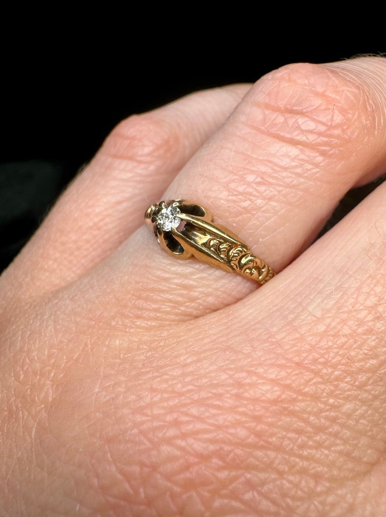 May include: A gold ring with a single diamond set in a decorative setting. The ring is worn on a finger.