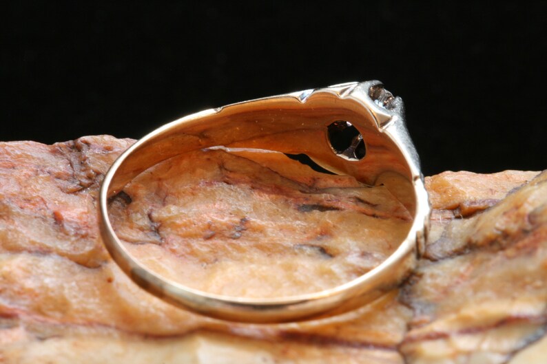 May include: A gold ring with a single diamond set in a delicate, ornate setting. The ring is shown from the underside, revealing the smooth, polished band.