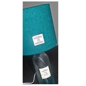 May include: A table lamp featuring a teal herringbone fabric lampshade with a Harris Tweed label and a clear, ribbed glass base made from an Isle of Harris Gin bottle with its original white label featuring black text.