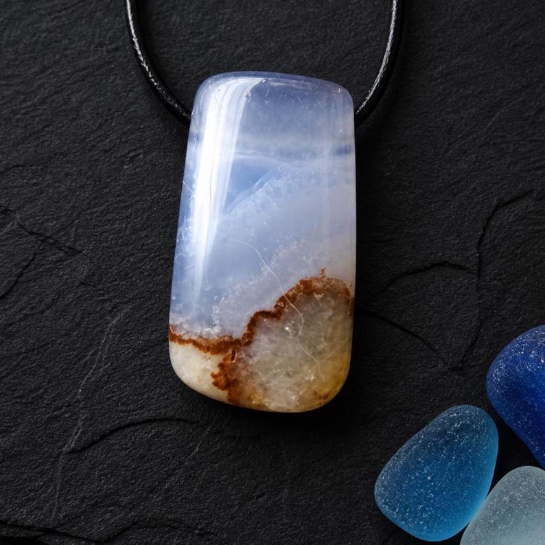 May include: A rectangular pendant necklace featuring a polished, translucent stone with a blue and brown pattern. The stone is suspended from a black cord, set against a dark background. Additional blue stones are in the background.