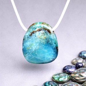 Può includere: Un ciondolo di pietra preziosa blu e marrone su un cordino bianco. Il ciondolo ha una superficie lucida e liscia con un mix di colori blu, turchese e marrone. Lo sfondo è di un colore neutro, con diverse pietre iridescenti.