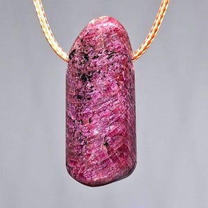 Könnte beinhalten: Ein lebendiger, länglicher, rosa Edelsteinanhänger mit strukturierter Oberfläche und dunklen Einschlüssen. Der Stein hängt an einer dünnen, hellbraunen Kordel. Der Anhänger hat eine natürliche, organische Form.