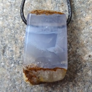 May include: A rectangular pendant with a blue and brown stone, suspended from a black cord. The stone has a translucent, layered appearance with shades of blue and brown. The pendant is likely a piece of jewelry.