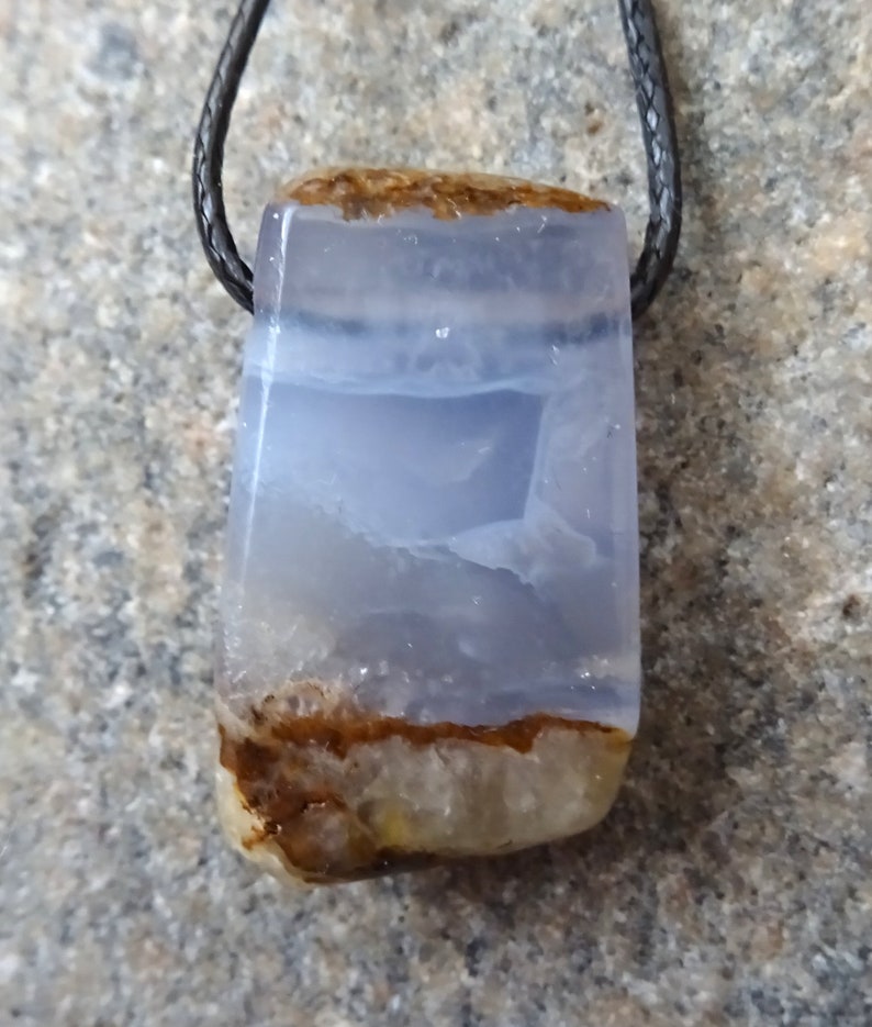 May include: A rectangular pendant with a blue and brown stone, suspended from a black cord. The stone has a translucent, layered appearance with shades of blue and brown. The pendant is likely a piece of jewelry.