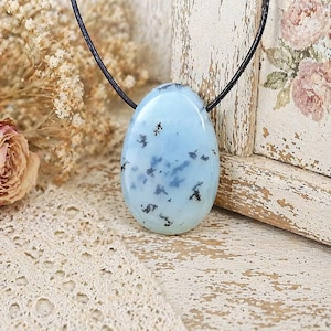 Può includere: Una collana con pendente ovale azzurro con inclusioni nere. La pietra è sospesa a un cordino nero. Il gioiello è esposto su una superficie di legno chiaro e rustico. Un gioiello elegante.