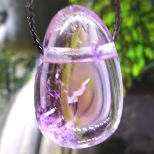 Könnte beinhalten: Ein violetter und transparenter Kristallanhänger an einer schwarzen Schnurkette. Der Kristall ist eiförmig und durchscheinend.