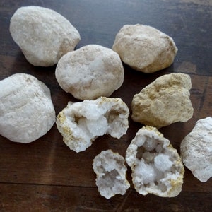 Könnte beinhalten: Eine Sammlung von acht Geoden, von denen einige aufgebrochen sind, um weiße Kristallformationen im Inneren zu zeigen. Die Geoden sind in verschiedenen Brauntönen und Weißtönen gehalten.