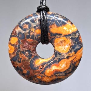 Puede incluir: Colgante en forma de rosquilla con cordón negro. El colgante presenta un patrón único de tonos naranja, azul oscuro y marrón. La piedra tiene un acabado pulido y un agujero en el centro, ideal para llevar como collar.