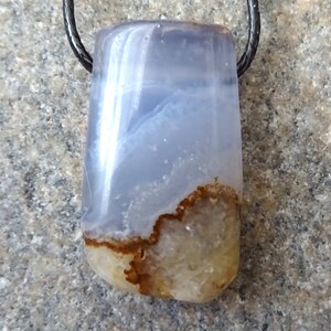 May include: A rectangular pendant made of polished blue and brown agate, suspended from a black cord. The stone features a translucent blue upper section and a lower section with brown and beige patterns. The pendant is likely a piece of jewelry.