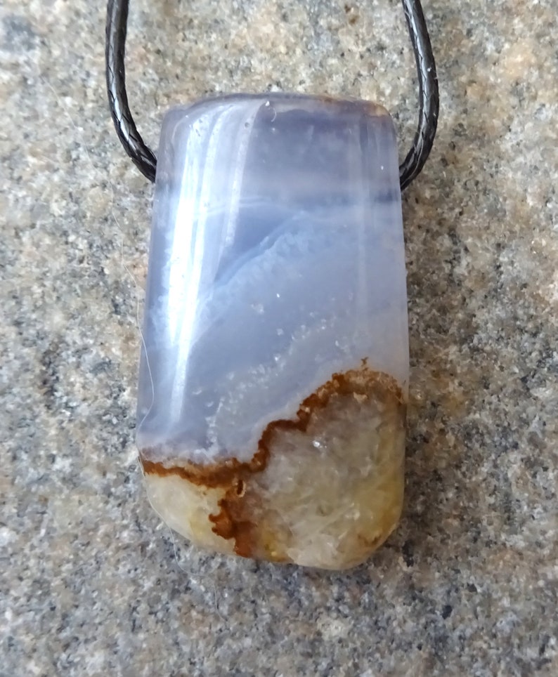 May include: A rectangular pendant made of polished blue and brown agate, suspended from a black cord. The stone features a translucent blue upper section and a lower section with brown and beige patterns. The pendant is likely a piece of jewelry.