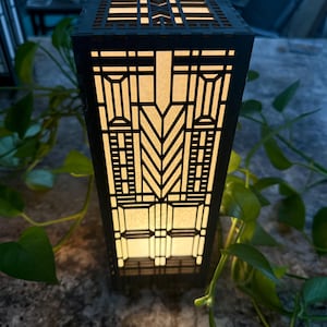 May include: A rectangular black lantern with a geometric design, illuminated from within. The lantern's intricate cutouts create a pattern of light and shadow. The design is reminiscent of art deco style. The lantern is surrounded by green foliage.