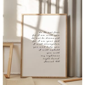May include: A framed print with a light wood frame displays the text "So do not fear, for I am with you...Isaiah 41:10" in elegant black script. The print is set against a neutral background.