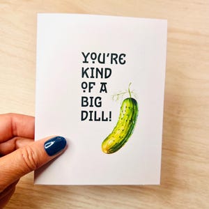 Peut inclure: Une carte blanche avec du texte noir qui dit "You're kind of a big dill!" et une illustration de cornichon vert.