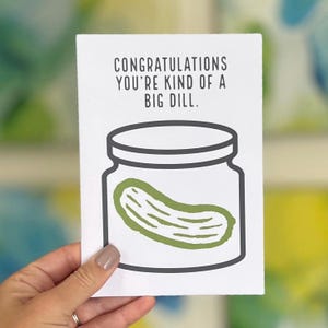 Peut inclure: Carte de vœux blanche tenue par une main, avec le texte "CONGRATULATIONS YOU'RE KIND OF A BIG DILL." En dessous, un dessin d'un bocal à cornichons avec un cornichon vert. Le design de la carte est simple et épuré.