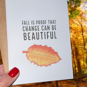 Puede incluir: Una tarjeta de felicitación blanca con el texto "FALL IS PROOF THAT CHANGE CAN BE BEAUTIFUL" y una ilustración de acuarela de una hoja de otoño. La tarjeta se sostiene contra un fondo borroso de follaje otoñal.