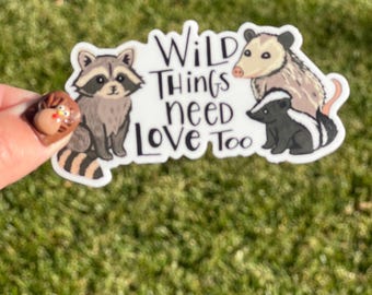 May include: A die-cut sticker featuring a gray raccoon, a tan opossum, and a black and white skunk. The text in the center reads Wild Things need LOVE Too in black lettering. The sticker has a white border and a glossy finish.