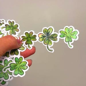 May include: A collection of green four-leaf clover stickers with white borders. The clovers have a watercolor effect, with varying shades of green and yellow. Some stickers are held in a hand against a neutral background.