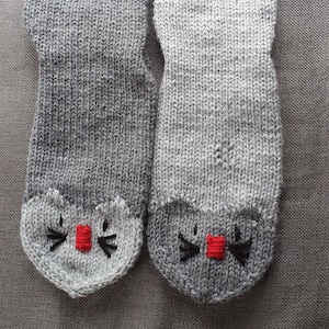 Könnte beinhalten: Ein Paar graue Stricksocken mit roten und schwarzen gestickten Katzenköpfen an den Zehen.