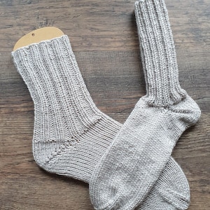 Könnte beinhalten: Ein Paar hellgraue Strickstrümpfe, die auf einer Holzoberfläche liegen. Die Socken haben ein Rippenmuster an Bein und Fuß, mit einer glatten Spitze und Ferse. Die Socken sind aus einem weichen, warmen Material gefertigt.