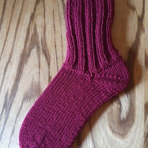 May include: A deep magenta knitted sock featuring a ribbed cuff and a smooth knit foot section. The sock is displayed on a cardboard insert, showcasing the vertical ribbing pattern extending from the top down to the heel and the solid knit texture.