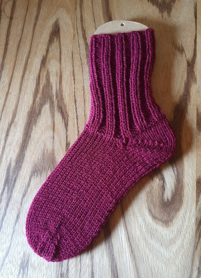 May include: A deep magenta knitted sock featuring a ribbed cuff and a smooth knit foot section. The sock is displayed on a cardboard insert, showcasing the vertical ribbing pattern extending from the top down to the heel and the solid knit texture.