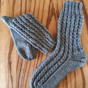 Può includere: Un paio di calzini di lana grigi con un motivo a treccia. Un calzino è piegato per mostrare il dettaglio della maglia.