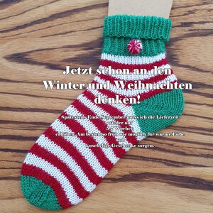 May include: A hand-knitted sock featuring horizontal red and white stripes with a green heel and toe. The cuff is green with a small red and white circular button. Text on the sock reads: Jetzt schon an den Winter und Weihnachten denken!