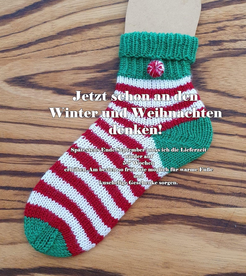May include: A hand-knitted sock featuring horizontal red and white stripes with a green heel and toe. The cuff is green with a small red and white circular button. Text on the sock reads: Jetzt schon an den Winter und Weihnachten denken!