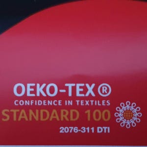 May include: A red rectangular label featuring the white text OEKO-TEX CONFIDENCE IN TEXTILES, the gold text STANDARD 100, the white code 2076-311 DTI, and a white circular graphic icon composed of interconnected loops on the right side.