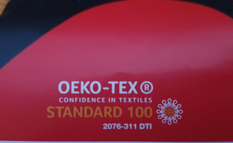 May include: A red rectangular label featuring the white text OEKO-TEX CONFIDENCE IN TEXTILES, the gold text STANDARD 100, the white code 2076-311 DTI, and a white circular graphic icon composed of interconnected loops on the right side.