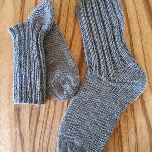 May include: A pair of hand-knitted grey socks featuring a ribbed cuff and a smooth knit foot section. The socks are crafted from a textured yarn with subtle variations in the grey tone, showcasing a classic construction with a defined heel.