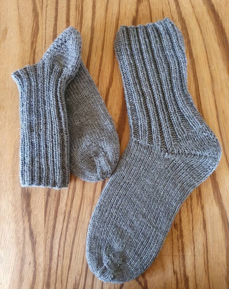 May include: A pair of hand-knitted grey socks featuring a ribbed cuff and a smooth knit foot section. The socks are crafted from a textured yarn with subtle variations in the grey tone, showcasing a classic construction with a defined heel.