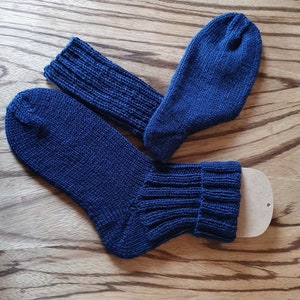 May include: A pair of dark blue knitted socks featuring a ribbed cuff design. One sock is laid flat while the other is positioned at an angle, with a small beige cardboard insert visible inside the cuff of the lower sock to maintain its shape.