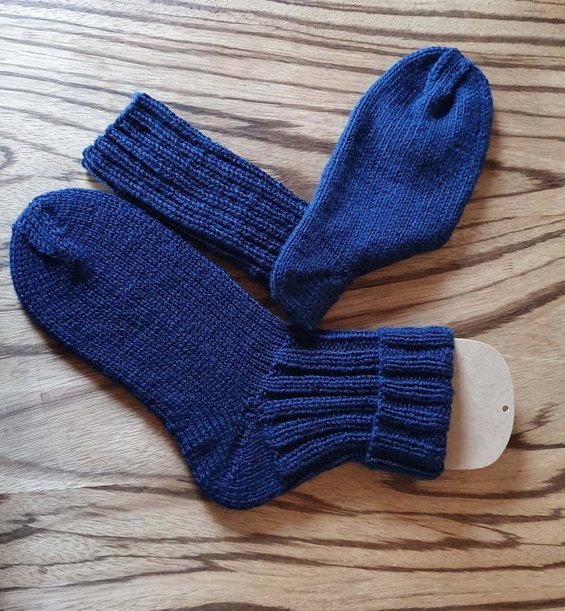 May include: A pair of dark blue knitted socks featuring a ribbed cuff design. One sock is laid flat while the other is positioned at an angle, with a small beige cardboard insert visible inside the cuff of the lower sock to maintain its shape.