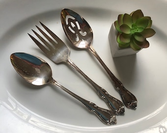 Könnte beinhalten: Drei silberne Besteckteile auf einem weißen Teller. Eine Gabel, ein Löffel und ein Schöpflöffel. Der Schöpflöffel hat ein dekoratives Design.