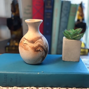 Könnte beinhalten: Eine kleine, weiße Keramikvase mit braunen und braunen Marmorierungen. Die Vase steht auf einem blauen Buch mit dem sichtbaren Titel "Dame". Eine kleine Sukkulente in einem grauen Topf steht neben der Vase.