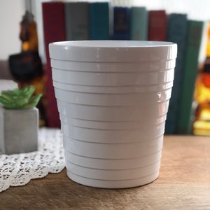 May include: White ceramic planter with a ribbed design. The planter is empty and sitting on a wooden surface.