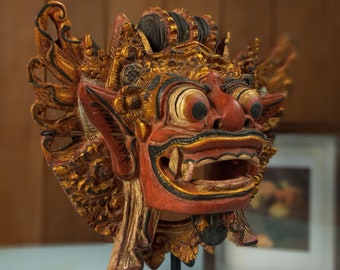 Può includere: Una maschera di legno con intagli intricati e vernice dorata. La maschera raffigura una creatura mitologica con un'espressione feroce, occhi grandi e una bocca spalancata. La maschera è montata su un supporto nero.