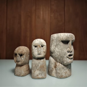 Puede incluir: Tres esculturas de piedra desgastada de cabezas humanas, cada una con rasgos faciales distintos. Las esculturas varían en tamaño, mostrando la más grande un rostro detallado y la más pequeña una forma simplificada. El color es una mezcla de gris y beige.