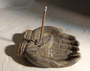 May include: A weathered, hand-shaped incense holder in a muted green-brown hue. The holder features intricate floral and leaf carvings, with a single incense stick inserted. The design evokes a sense of tranquility and is suitable for home decor.