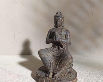 May include: A bronze-colored Buddha statue in a seated meditation pose. The figure has hands pressed together in prayer, wearing a draped robe, and is seated on a detailed circular base. The statue is on a white surface with a textured background.