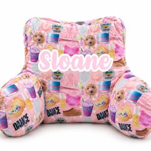 May include: A pink reading pillow featuring a repeating pattern of puppies, colorful drinks, nail polish bottles, and the word DANCE. The name Sloane is printed in large white script across the center of the backrest portion.
