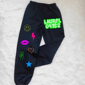 May include: Black sweatpants with a variety of colorful graphic designs, including a smiley face, lightning bolt, lips, heart, peace sign, and star. The text "LAUREL 04352" is printed in green on the right leg.