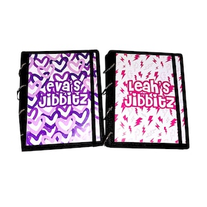 May include: Two black binders with colorful covers. One binder has a purple and pink heart pattern with the text "Eva's Jibbitz". The other binder has a white background with pink lightning bolts and the text "Leah's Jibbitz".