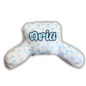 May include: A white reading pillow featuring a repeating pattern of light blue bows. The name Aria is embroidered in large, light blue, bubble-style letters with a black outline across the center of the backrest portion of the pillow.