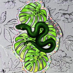 May include: A die-cut sticker featuring a dark green snake coiled over bright lime green monstera leaves. The sticker has a holographic glitter border and includes the text STONERADVISORYART printed in black along the right side of the design.