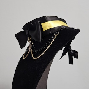 May include: Black choker necklace with a large bow and gold chain accents. The choker features a yellow ribbon detail and a small charm. The necklace is displayed on a black velvet form.