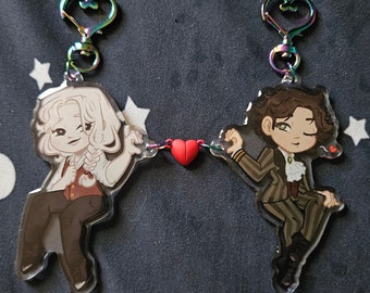 May include: Two cartoon character keychains connected by a red heart charm. The keychain on the left features a white-haired character with a brown jacket and black pants. The keychain on the right features a brown-haired character with a brown jacket and black pants. Both characters are smiling.