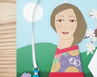 A 5x7 inch Flat Card with a unique paper person (not printed) in a kimono and tassel becoming a bookmark when removed from 2 photo corners.