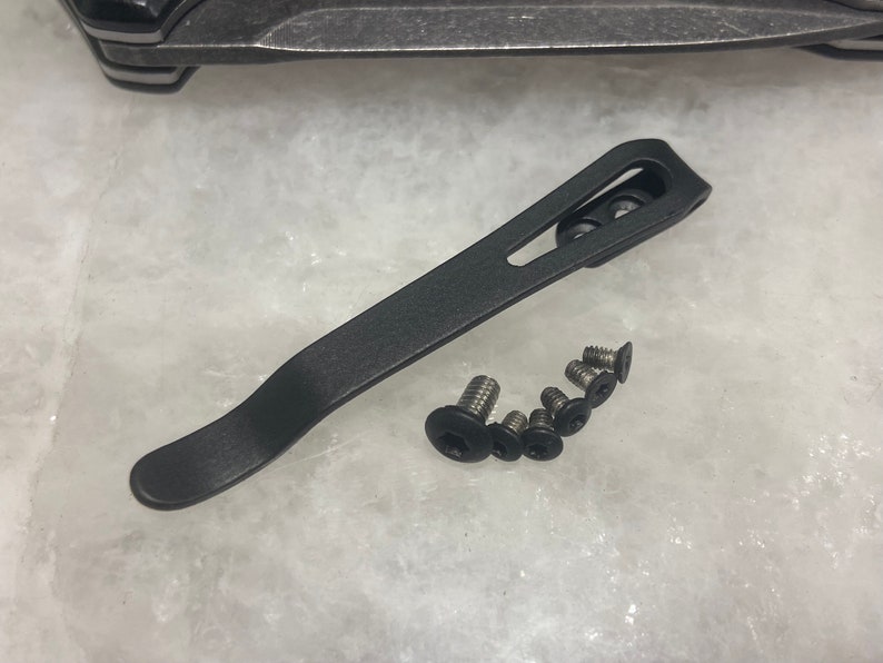 May include: A black metal pocket clip with a curved design. The clip is shown with six silver screws.